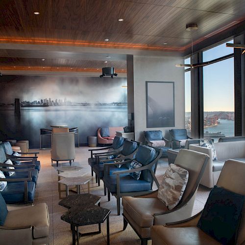 A modern lounge area with ample seating, large windows offering a waterfront view, and a cityscape mural on the back wall.