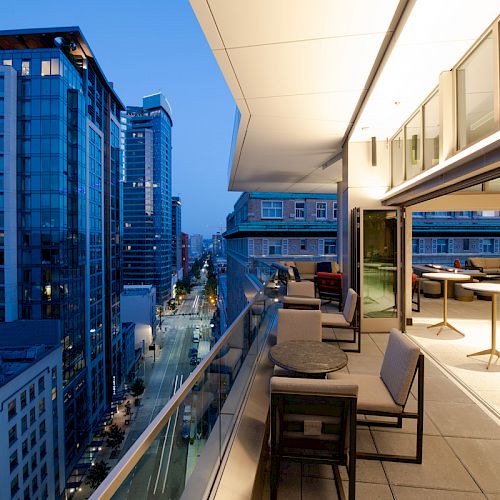 The image shows a modern cityscape at twilight with high-rise buildings and a view from a spacious, furnished balcony overlooking the street.