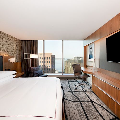 This image shows a modern hotel room with a large bed, a wall-mounted TV, a desk and chair, and a window offering a city view.