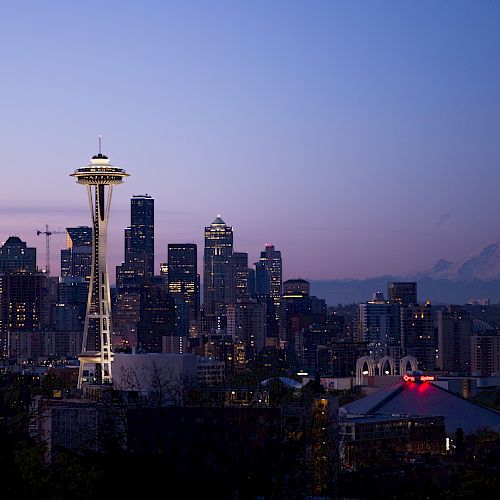The image shows the Seattle skyline at dusk with the Space Needle prominently visible, Mt. Rainier in the background, and various buildings lit up.