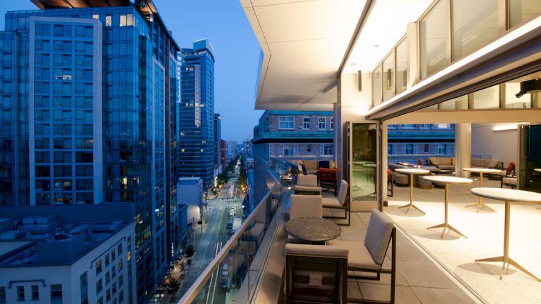 A modern, glass-walled balcony with seating overlooks a cityscape filled with lit-up high-rise buildings at dusk, offering an urban atmosphere.