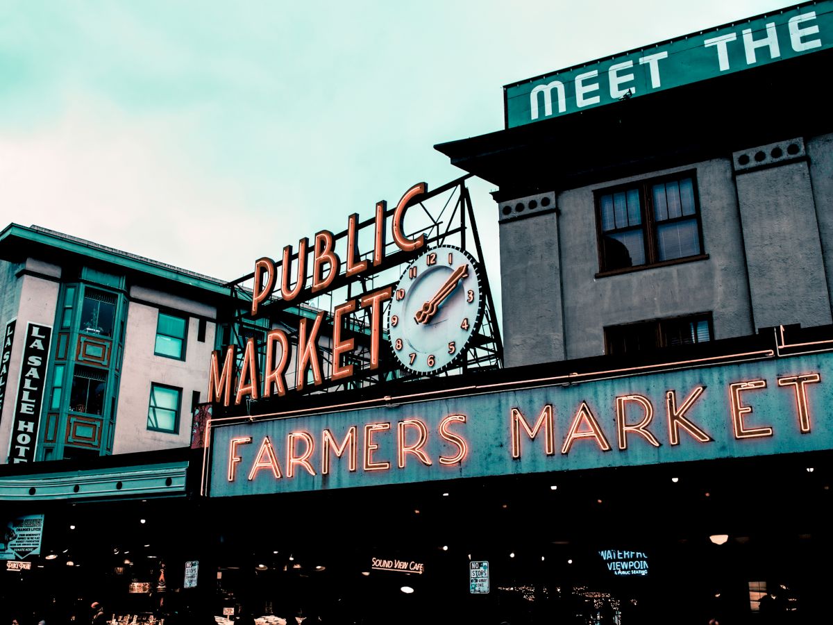 The image shows an iconic "Public Market" and "Farmers Market" sign with a clock above, located in a bustling urban area, likely during daytime.