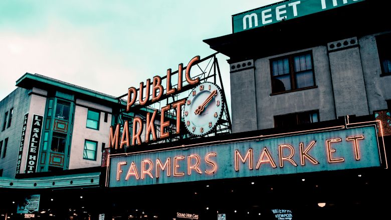 The image shows an iconic "Public Market" and "Farmers Market" sign with a clock above, located in a bustling urban area, likely during daytime.