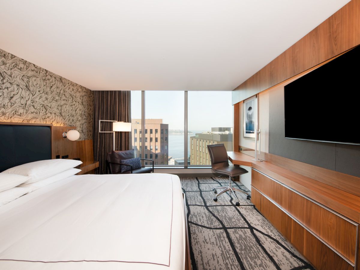 Modern hotel room with a large bed, flat-screen TV, desk, chair, and floor-to-ceiling window offering a city view over water.