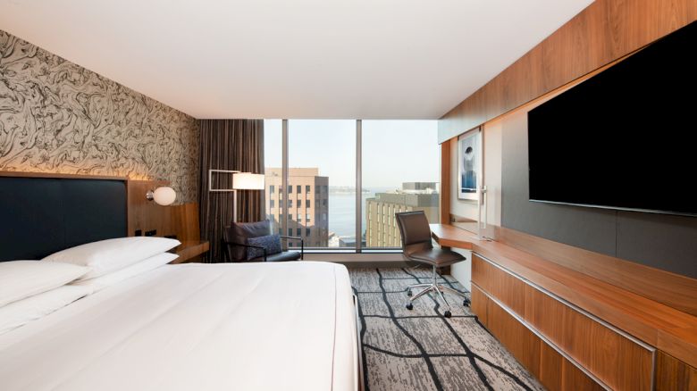 Modern hotel room with a large bed, flat-screen TV, desk, chair, and floor-to-ceiling window offering a city view over water.