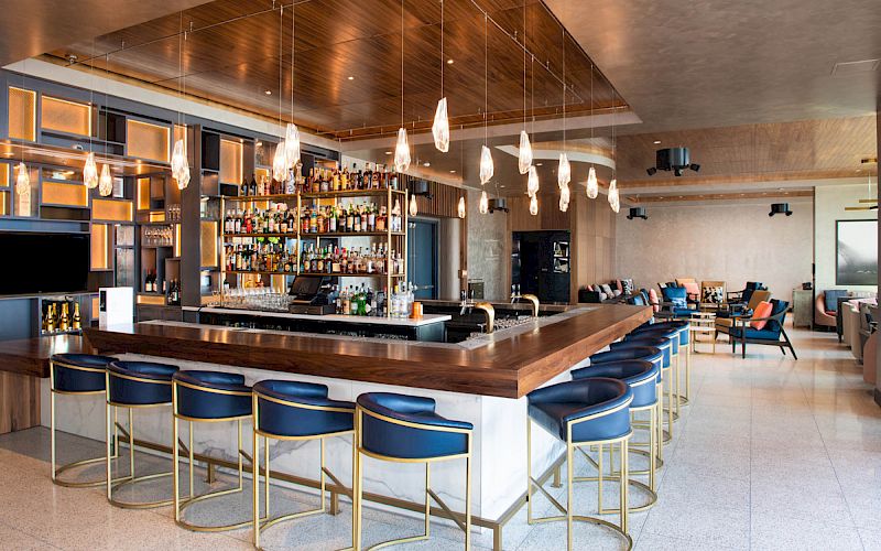 A modern bar scene with a well-stocked shelf, wooden counter, blue chairs, pendant lights, and a seating area with cushioned chairs and tables in the background.