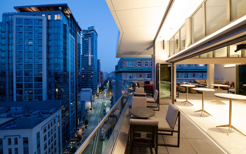 A modern cityscape view from an open balcony at dusk, featuring high-rise buildings, outdoor seating, and a well-lit interior space.