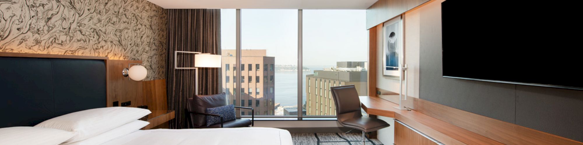A modern hotel room with a large bed, desk, chair, wall-mounted TV, and a window offering a city view.