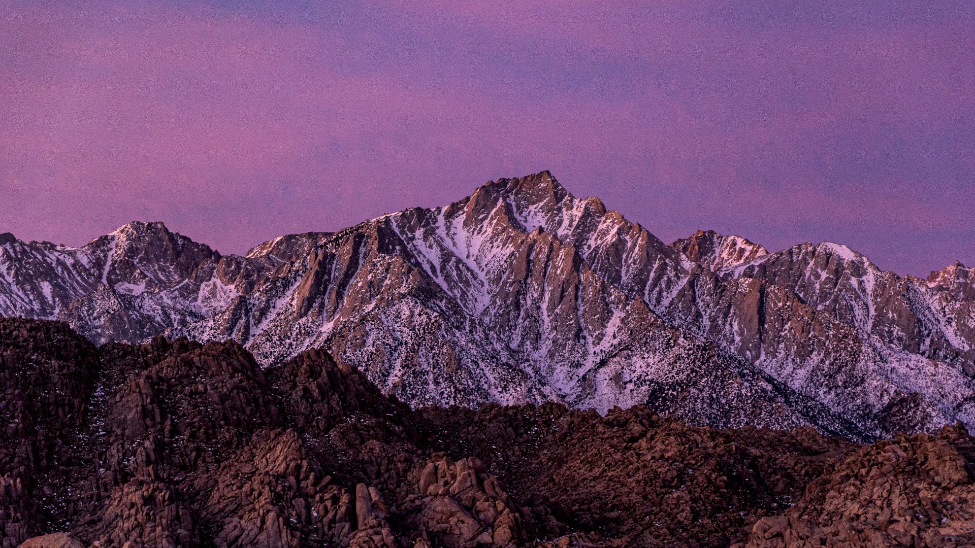 A mountain range with snow-capped peaks is under a purple sky, showing rugged terrain and beautiful snowy textures.