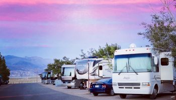 A line of motorhomes parked along a scenic road at sunset, with mountains in the distance and a colorful pink-purple sky.