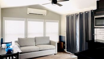 A modern living room with a grey sofa, white blinds, an air conditioner, dark curtains, a ceiling fan, and a kitchenette with dark cabinets and white tiles.