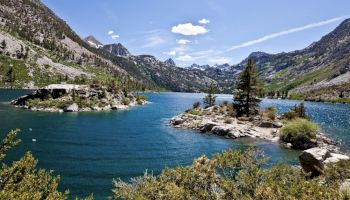 A pristine mountain lake surrounded by rugged mountains and scattered trees, with clear blue water under a bright, sunny sky with a few clouds.