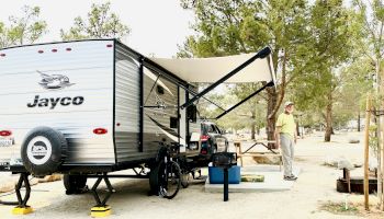 A person stands next to a Jayco travel trailer set up in a campsite with trees around, featuring an awning, outdoor gear, and a picnic table.
