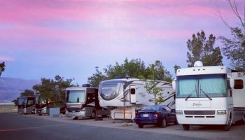 The image shows several RVs and a car parked under a pink and blue evening sky with trees and mountains in the background.