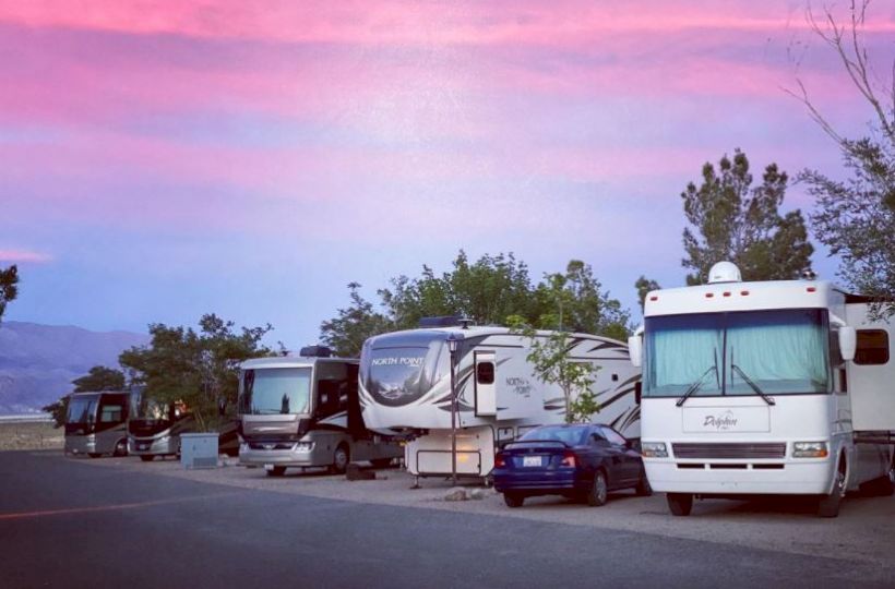 The image shows several RVs and a car parked under a pink and blue evening sky with trees and mountains in the background.