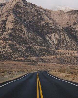 A straight, empty road leading towards rocky, mountainous terrain under a mostly overcast sky with patches of clear weather.