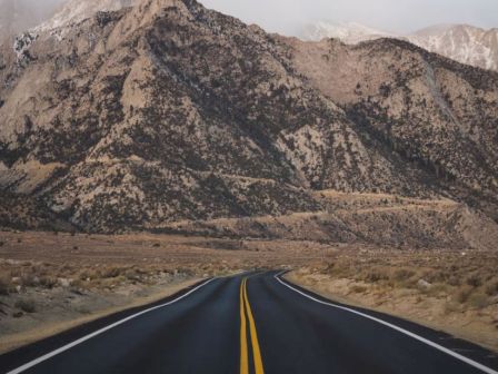 A straight, empty road leading towards rocky, mountainous terrain under a mostly overcast sky with patches of clear weather.