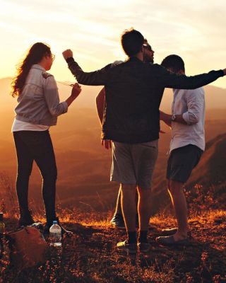 A group of people on a mountain enjoying a sunset; they are talking, gesturing and possibly dancing. The scenic view includes distant hills.