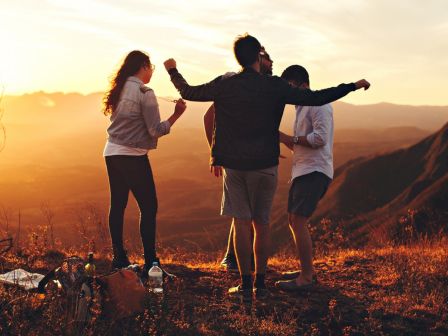 A group of people on a mountain enjoying a sunset; they are talking, gesturing and possibly dancing. The scenic view includes distant hills.