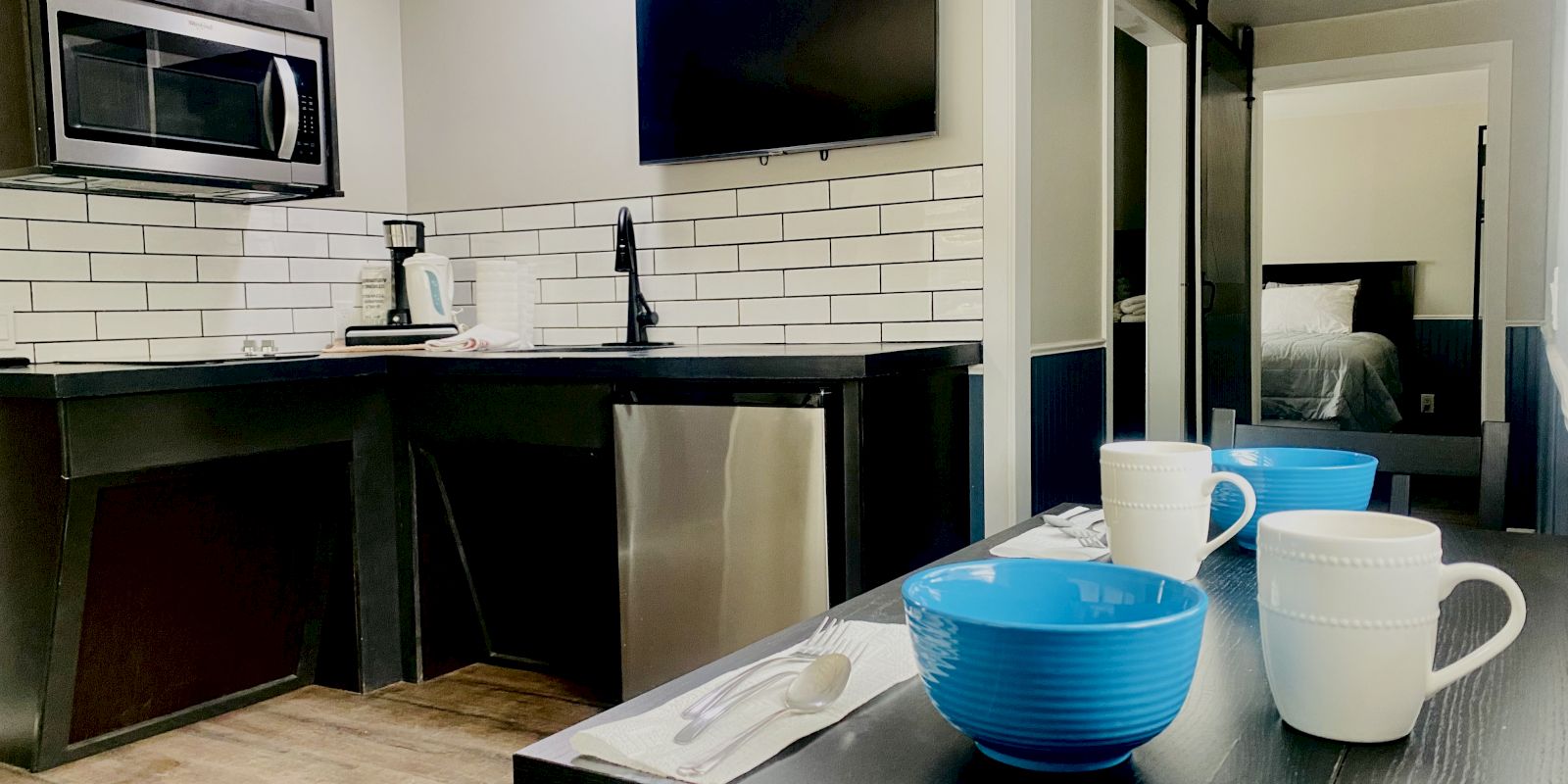 The image shows a modern kitchen with a microwave, sink, and TV, plus a dining area with a table set with bowls and cups. A bedroom is visible in the background.