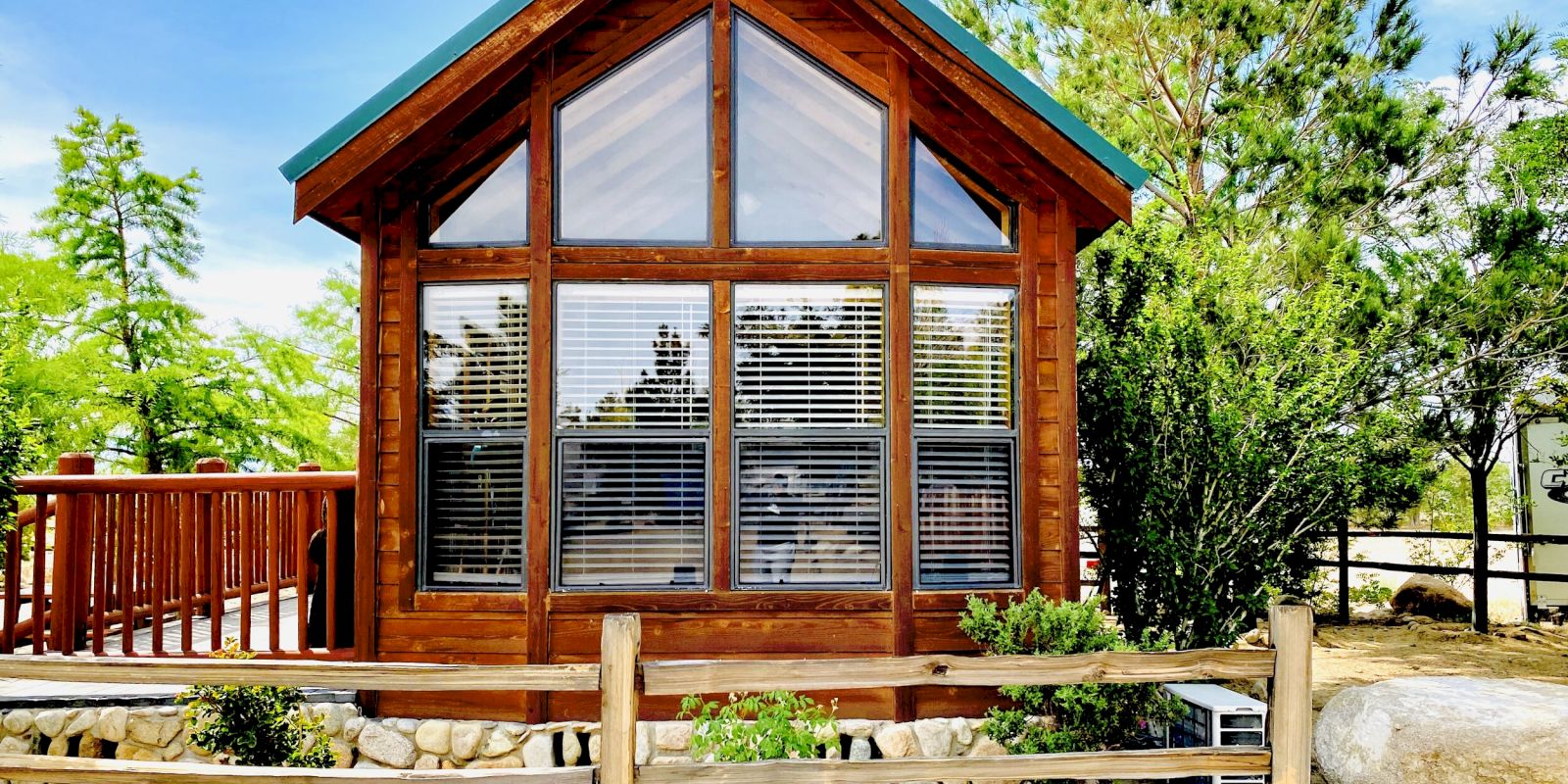 This image shows a small, charming wooden cabin with large windows and a deck, surrounded by trees and a fenced garden area, under a bright blue sky.