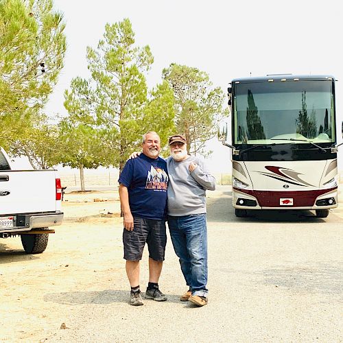 Two people standing in front of an RV, with a white truck to their left and a background of trees and open space.