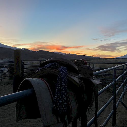 A sunset over a rural landscape with saddles hanging on a fence, and mountains in the background.
