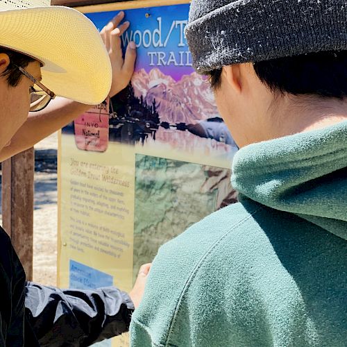 Two people are looking at an information board about "Trail/Wood." One is wearing a cowboy hat, and the other a beanie with a green hoodie.
