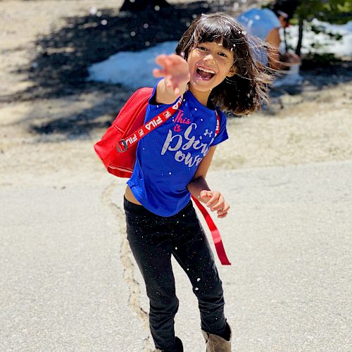 A child wearing a blue shirt and black pants with brown boots is smiling and waving outdoors on a sunny day. Snow and trees are in the background.