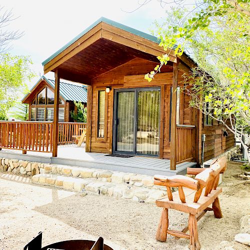 The image shows a cozy wooden cabin surrounded by nature, with a porch, a bench, and trees nearby, giving a peaceful, rustic feeling.