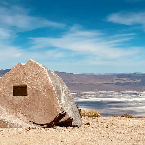 A large rock with a plaque is set against a vast landscape of mountains and a partly cloudy sky under which stretches a salt flat.