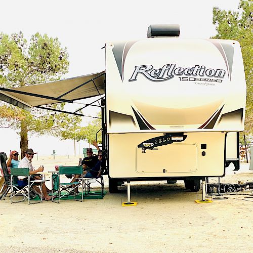 A group of people are sitting under the awning of an RV labeled “Reflection” in a campsite with trees surrounding them.