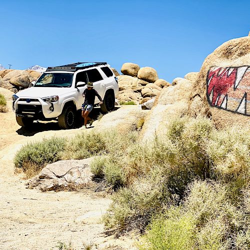 A white SUV is off-roading in a rocky, desert landscape with a person nearby; large rock painted with sharp teeth is visible.
