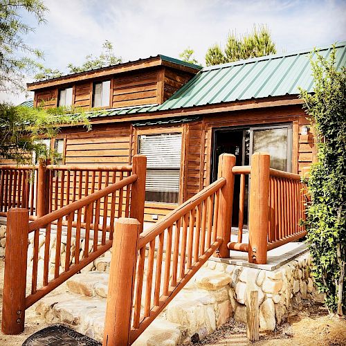 A cozy log cabin with green roofing and a front porch, set against a scenic backdrop of trees and a clear sky, featuring wooden railings and stone steps.