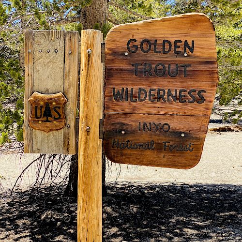 A wooden sign reads "Golden Trout Wilderness, Inyo." Beside it is a small USFS shield symbol attached to a post, with trees in the background.