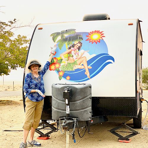 A person in casual clothing stands pointing at a trailer with a colorful mural of a woman in a hula outfit on the front, in an outdoor setting.