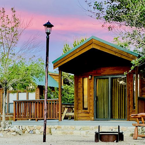 A wooden cabin with a porch is nestled among trees at sunset, featuring a nearby lamppost and a fire pit in the foreground.