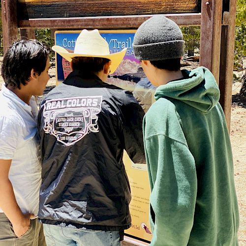 Three people are looking at an informational sign outdoors; one wears a cowboy hat, another a hoodie.