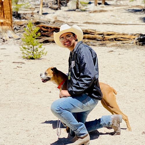 A person wearing a cowboy hat is kneeling on sandy terrain, smiling, and holding the leash of a tan and white dog. Trees and logs are in the background.