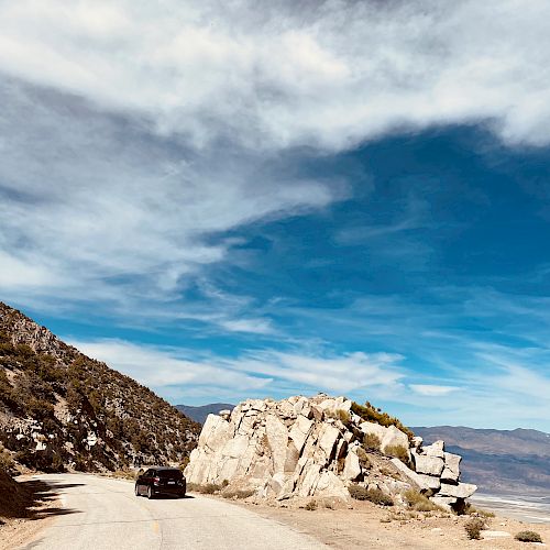 A scenic mountain road with a black car traveling near large rock formations under a partly cloudy sky. Blue mountains are visible in the distance.