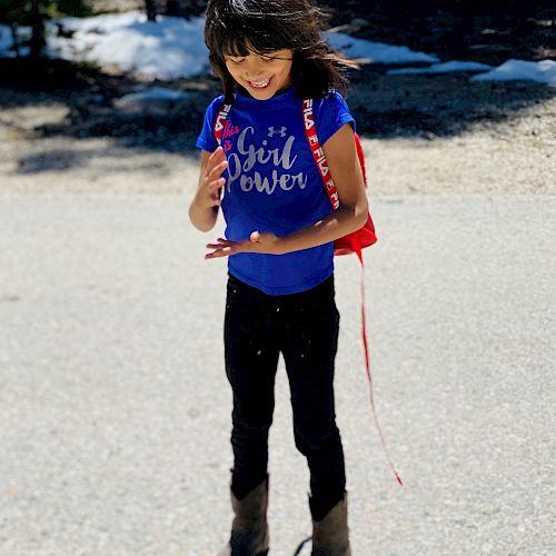 A young girl wearing a "Girl Power" shirt, jeans, and boots stands outdoors while holding something, with trees and patches of snow in the background.