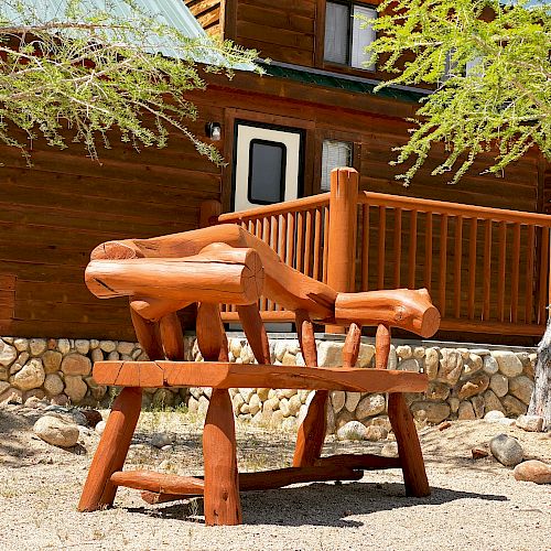 A rustic wooden bench is positioned between two trees in front of a log cabin with a stone foundation and a wooden deck.