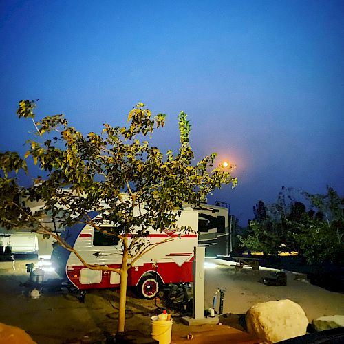 A small camper trailer parked near a tree at dusk with lights illuminating the area and a car in the background.