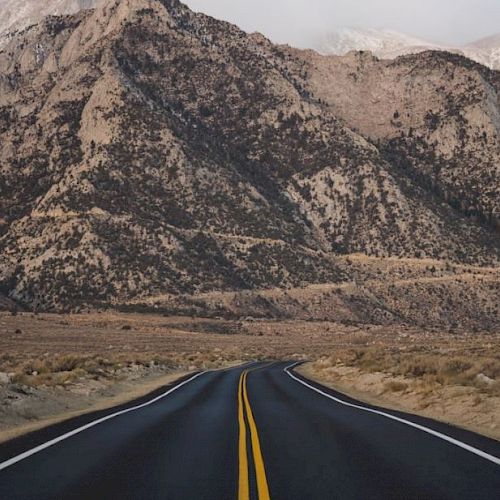 An empty, paved road stretches forward towards a rugged mountain range with sparse, arid landscape on either side and a cloudy sky above.