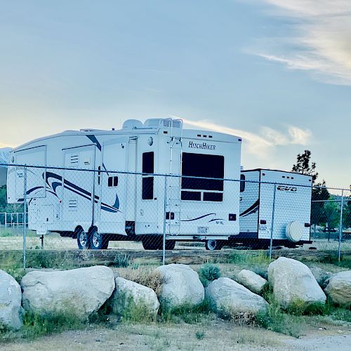 The image shows a white recreational vehicle (RV) parked behind a chain-link fence, with large rocks in the foreground and a scenic sky in the background.