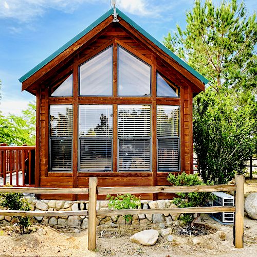 A small, wooden cabin with large windows and a green roof sits in a natural setting with trees, a stone base, and a wooden fence surrounding it.