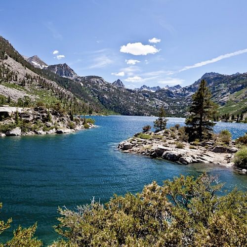 A serene lake surrounded by mountains with clear blue water, islands with trees, and a vibrant blue sky with some clouds completes the scenic view.