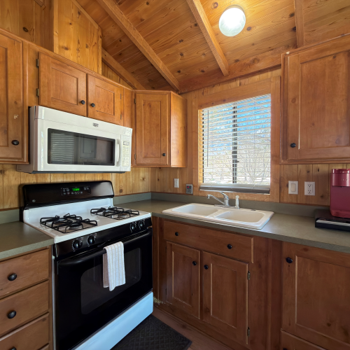 A cozy wood-paneled kitchen with a white microwave, gas stove, green countertops, and a window over a sink, plus a red coffee maker.