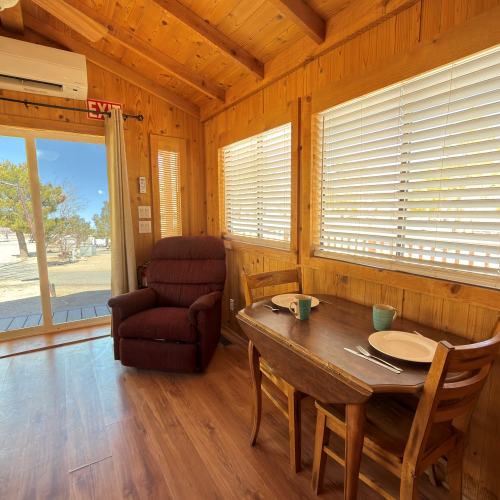 Cozy wood-paneled cabin interior with a small dining table, two chairs, a red recliner, large windows, and a sliding door to a sunny outdoor view.