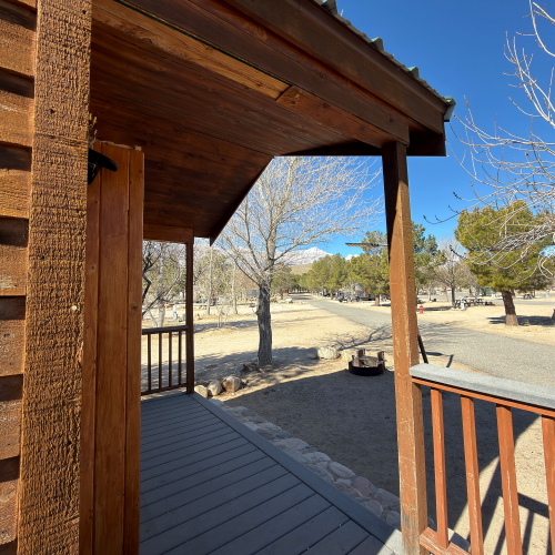 A wooden cabin porch with a railing overlooks a dry, sparsely treed yard under a clear blue sky; bare trees and distant hills create a quiet rural scene.